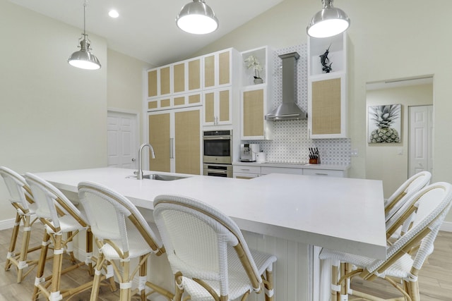 kitchen with double oven, a sink, wall chimney range hood, backsplash, and light wood finished floors