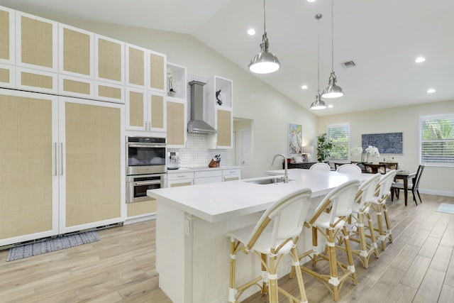 kitchen featuring double oven, a kitchen island with sink, light wood-style flooring, a sink, and wall chimney range hood