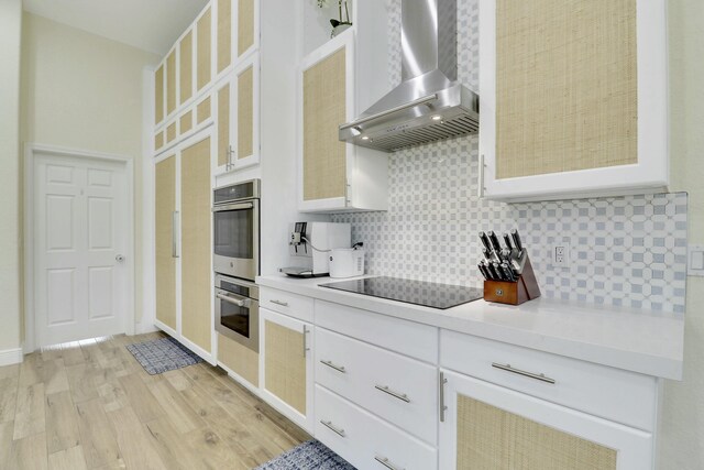 kitchen featuring black electric cooktop, light countertops, decorative backsplash, wall chimney exhaust hood, and light wood finished floors