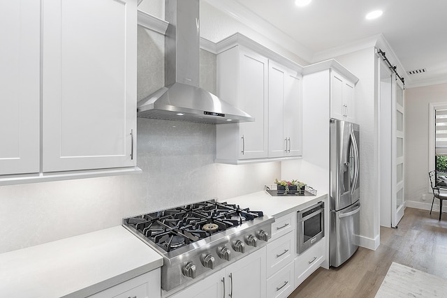 kitchen with wall chimney range hood, light countertops, a barn door, ornamental molding, and stainless steel appliances