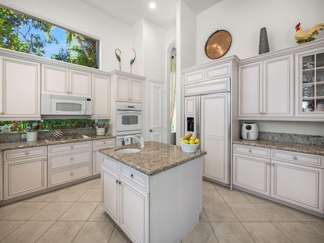 kitchen featuring an island with sink, a sink, light stone counters, white appliances, and light tile patterned flooring