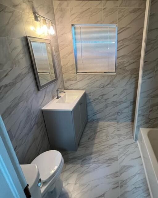 full bathroom with tile walls, toilet, a tub to relax in, marble finish floor, and vanity