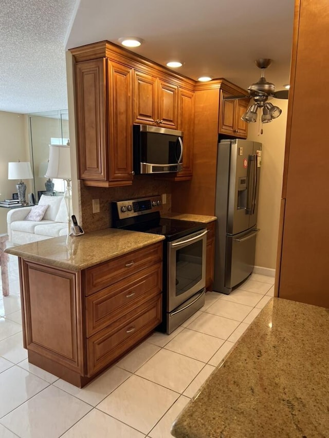 kitchen with brown cabinetry, ceiling fan, light stone counters, appliances with stainless steel finishes, and light tile patterned flooring