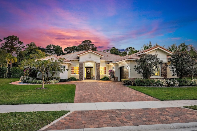 mediterranean / spanish home featuring a tile roof, decorative driveway, a lawn, and stone siding