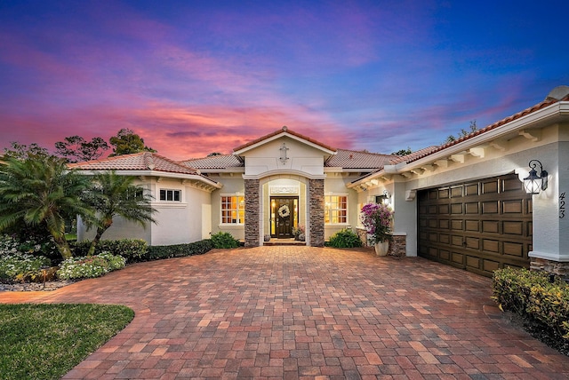 mediterranean / spanish-style home featuring stucco siding, decorative driveway, stone siding, an attached garage, and a tiled roof