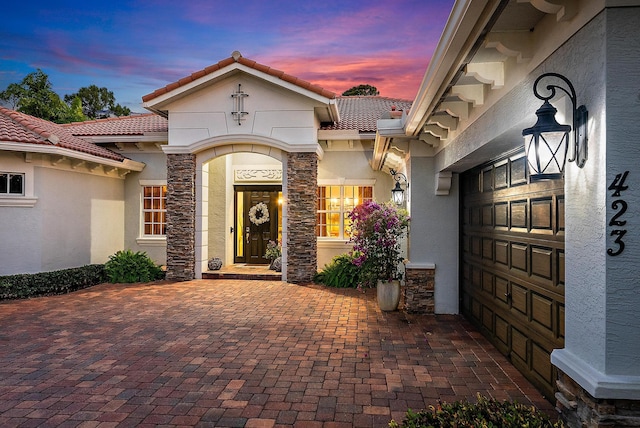 exterior entry at dusk featuring a tile roof, stone siding, and stucco siding