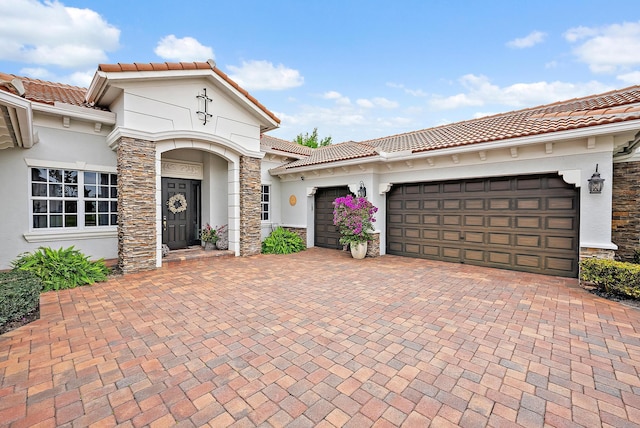 mediterranean / spanish home featuring an attached garage, stucco siding, stone siding, a tile roof, and decorative driveway