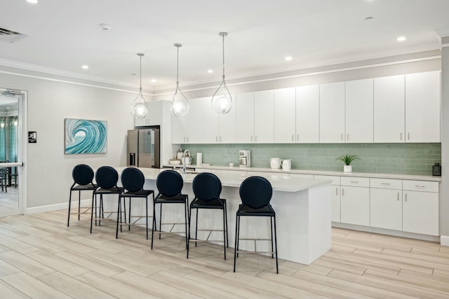 kitchen with backsplash, crown molding, light countertops, stainless steel refrigerator with ice dispenser, and a kitchen island with sink