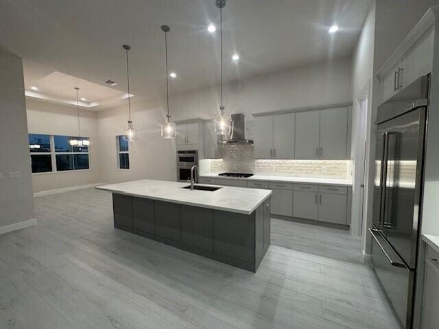 kitchen with gray cabinetry, wall chimney range hood, a tray ceiling, built in fridge, and a sink