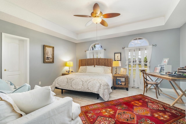 bedroom with a raised ceiling, an office area, light wood-style flooring, and ceiling fan