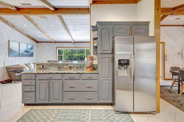 kitchen with light tile patterned floors, lofted ceiling with beams, gray cabinetry, stainless steel refrigerator with ice dispenser, and wood walls