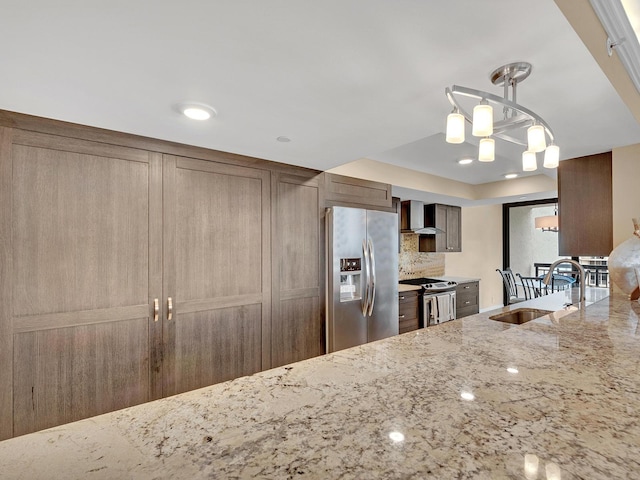 kitchen featuring a sink, light stone counters, backsplash, stainless steel appliances, and wall chimney range hood