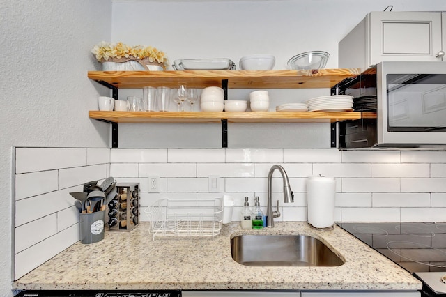 kitchen with open shelves, stainless steel microwave, light stone counters, backsplash, and a sink