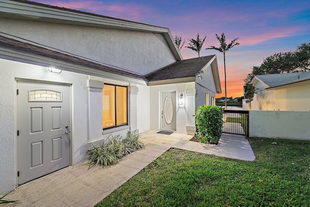 exterior entry at dusk featuring stucco siding, a lawn, and a gate