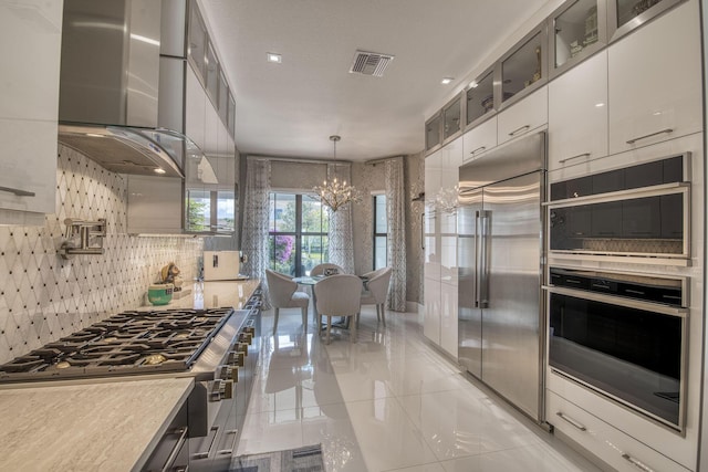 kitchen featuring backsplash, appliances with stainless steel finishes, wall chimney range hood, glass insert cabinets, and a chandelier