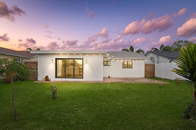 back of house at dusk with stucco siding, fence, a lawn, and a gate