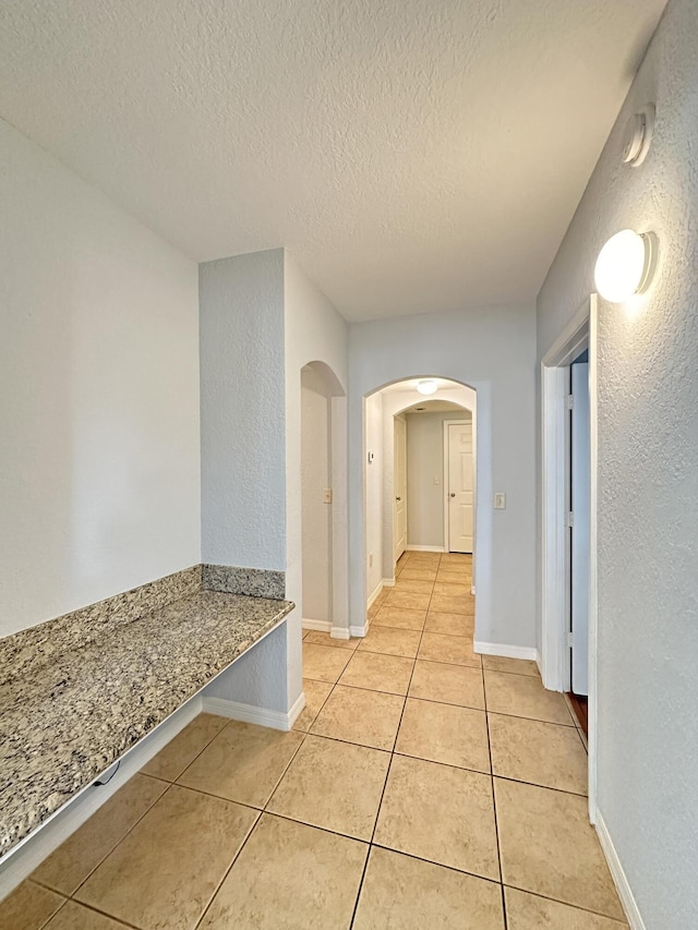 hall featuring arched walkways, light tile patterned floors, a textured ceiling, and baseboards