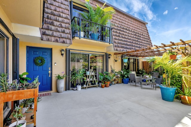 view of exterior entry featuring stucco siding, a pergola, mansard roof, a balcony, and a patio area