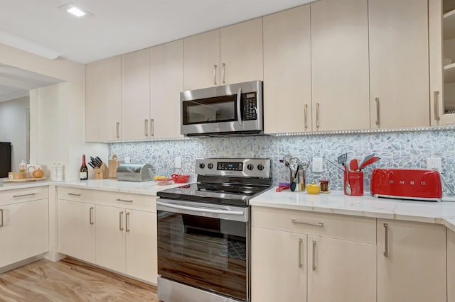 kitchen with light wood-type flooring, cream cabinetry, light stone counters, tasteful backsplash, and appliances with stainless steel finishes