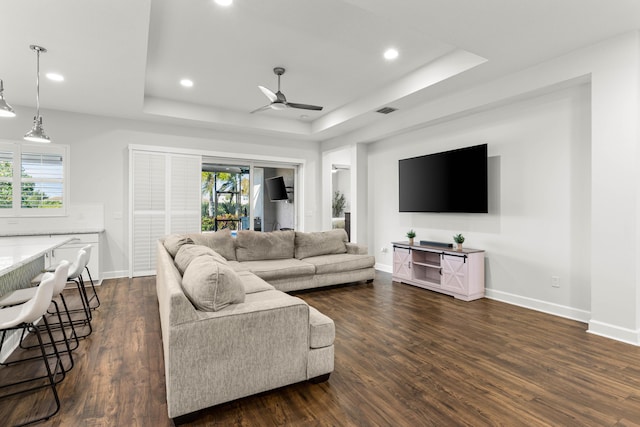 living area featuring a raised ceiling, baseboards, dark wood-type flooring, and ceiling fan