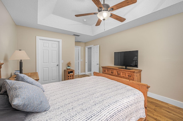 bedroom featuring a ceiling fan, wood finished floors, baseboards, a tray ceiling, and a textured ceiling