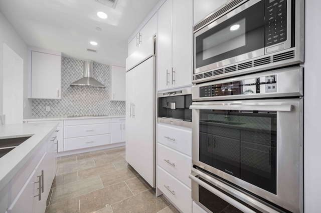 kitchen with white cabinetry, built in appliances, backsplash, and wall chimney range hood