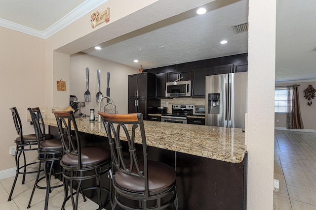 kitchen featuring dark cabinetry, light stone counters, ornamental molding, appliances with stainless steel finishes, and tasteful backsplash