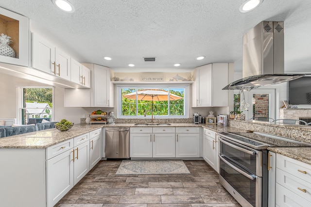 kitchen with a peninsula, island exhaust hood, stainless steel appliances, white cabinetry, and a sink