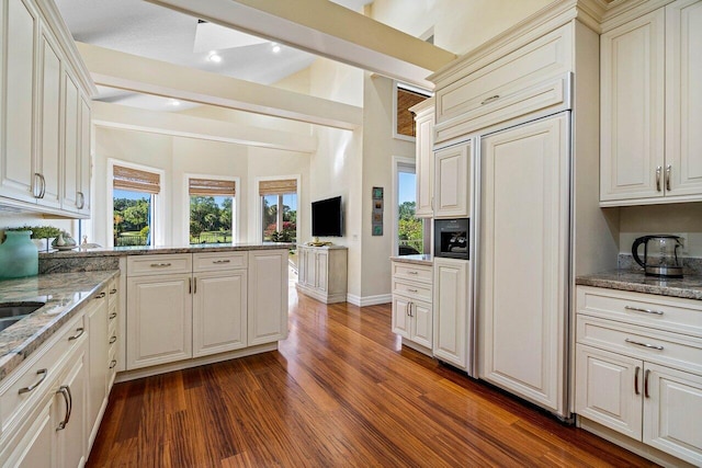 kitchen featuring beam ceiling, dark wood-type flooring, a peninsula, light stone countertops, and paneled built in refrigerator