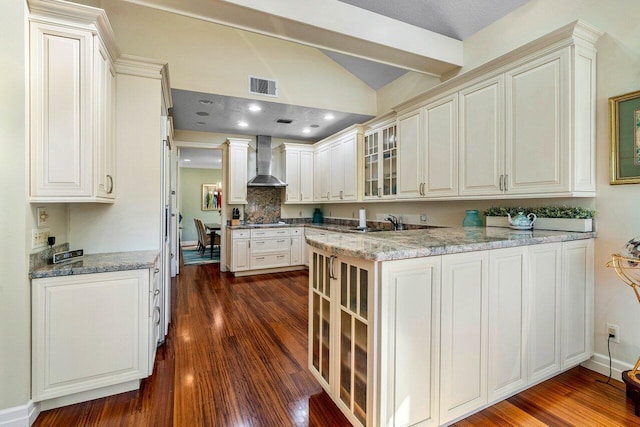 kitchen with visible vents, wall chimney range hood, glass insert cabinets, dark wood-style flooring, and vaulted ceiling