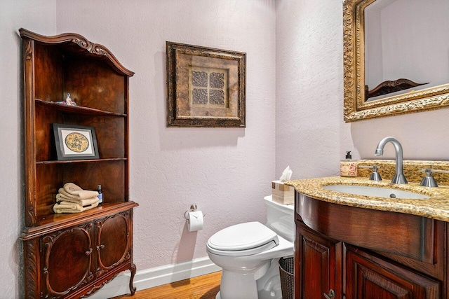 half bathroom featuring baseboards, toilet, wood finished floors, a textured wall, and vanity