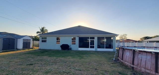 back of property at dusk featuring fence, a yard, a sunroom, stucco siding, and a storage unit