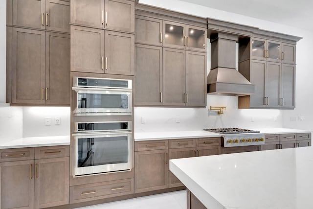 kitchen with glass insert cabinets, stainless steel appliances, light countertops, and wall chimney range hood