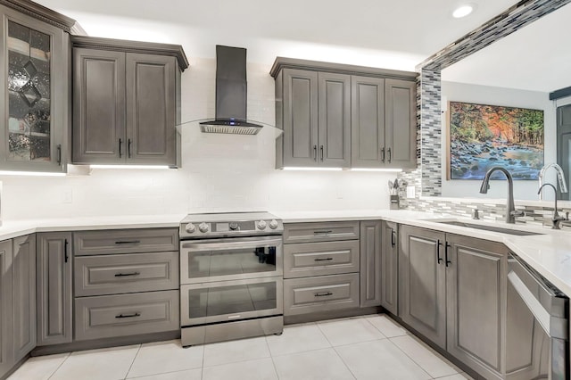 kitchen featuring gray cabinetry, glass insert cabinets, wall chimney range hood, stainless steel appliances, and a sink