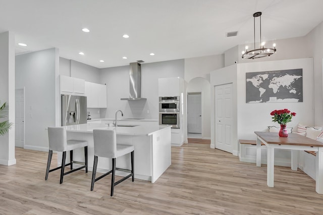 kitchen featuring a center island with sink, arched walkways, stainless steel appliances, light countertops, and wall chimney exhaust hood