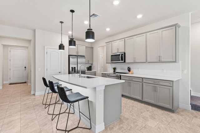 kitchen featuring gray cabinetry, a kitchen island with sink, a sink, appliances with stainless steel finishes, and a breakfast bar area