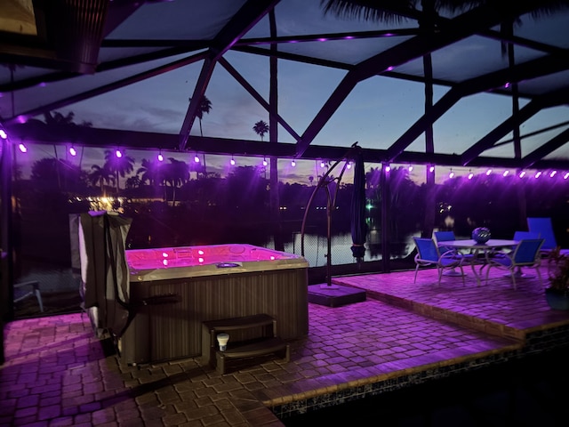 patio at twilight featuring a lanai, a water view, and a hot tub