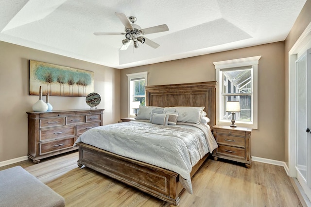 bedroom with ceiling fan, baseboards, light wood-type flooring, a tray ceiling, and a textured ceiling