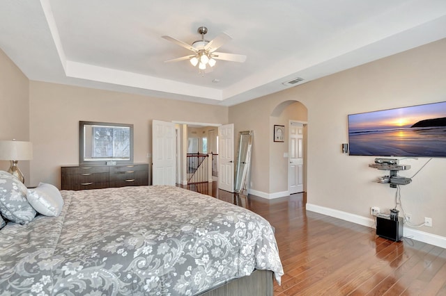 bedroom with a tray ceiling, wood finished floors, arched walkways, and baseboards