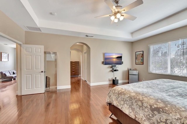 bedroom with visible vents, baseboards, light wood-type flooring, a tray ceiling, and arched walkways