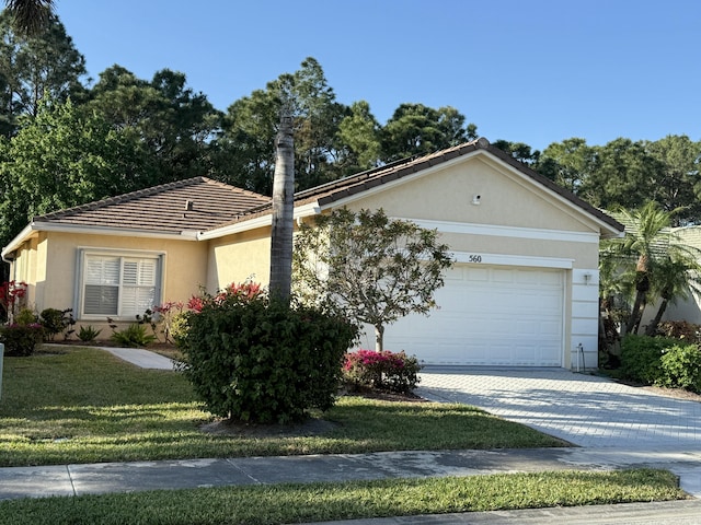 ranch-style home with stucco siding, decorative driveway, a front yard, an attached garage, and a tiled roof