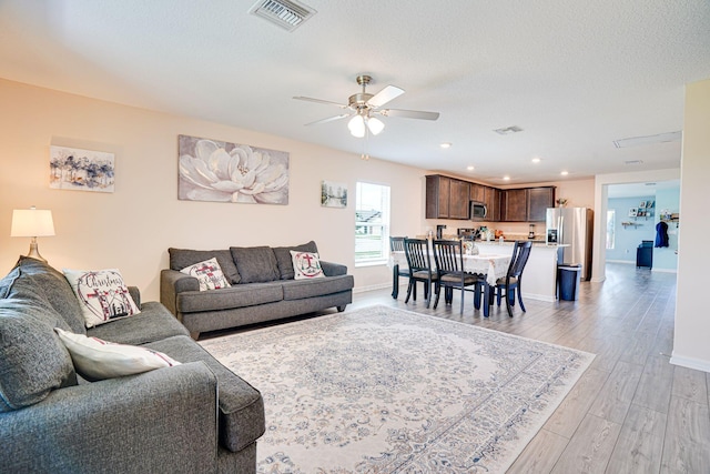 living room featuring light wood finished floors, visible vents, a textured ceiling, and a ceiling fan