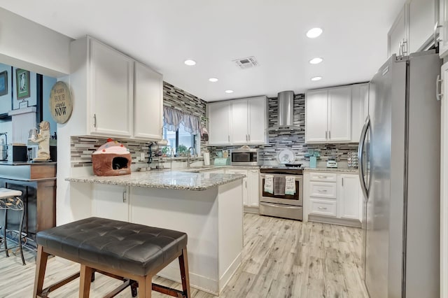 kitchen featuring light stone counters, a peninsula, light wood-style floors, stainless steel appliances, and wall chimney exhaust hood