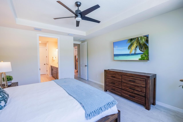 bedroom featuring a ceiling fan, baseboards, light wood finished floors, a tray ceiling, and ensuite bathroom