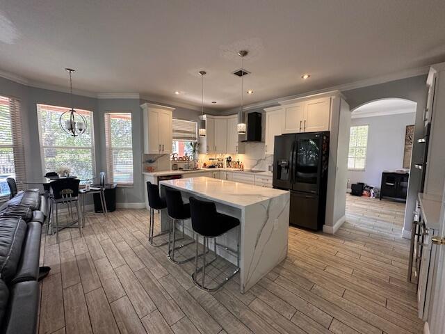 kitchen with tasteful backsplash, light wood finished floors, ornamental molding, black refrigerator with ice dispenser, and wall chimney exhaust hood