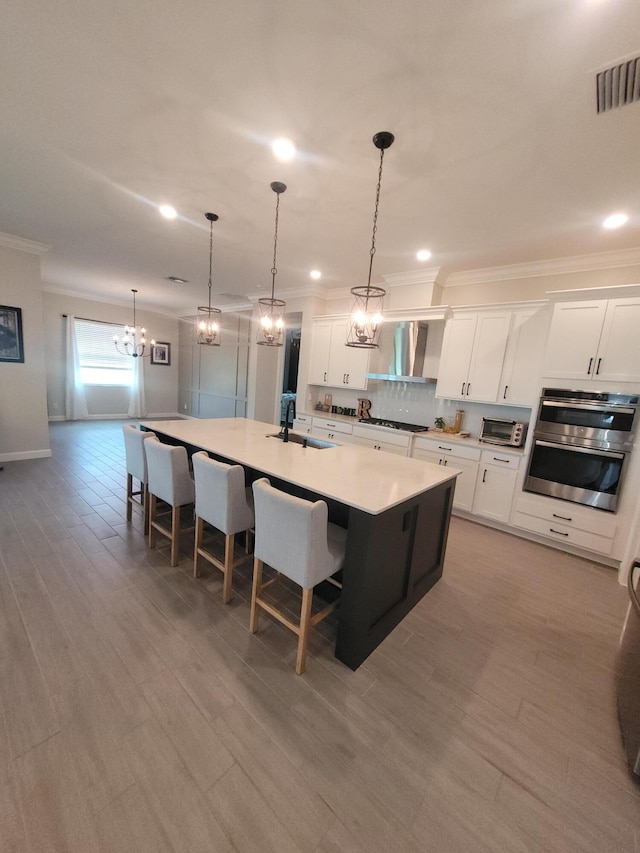 kitchen featuring visible vents, a kitchen island with sink, crown molding, wall chimney range hood, and a notable chandelier