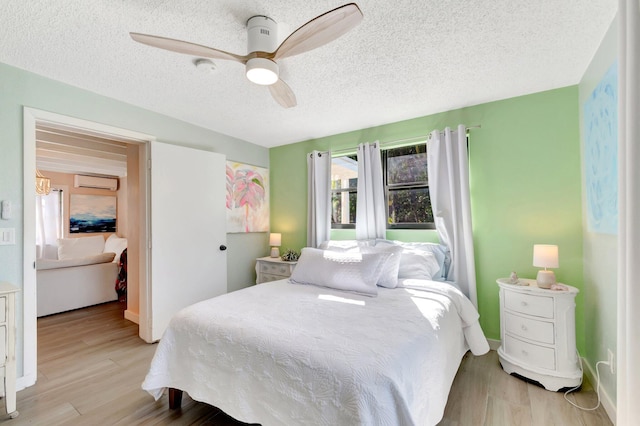 bedroom featuring light wood finished floors, a textured ceiling, a wall mounted air conditioner, and ceiling fan