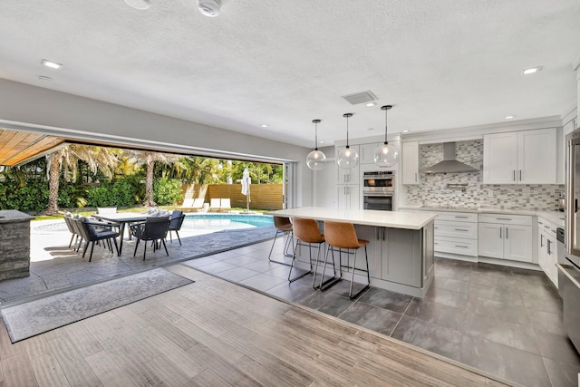 kitchen with backsplash, a kitchen island, double oven, light countertops, and wall chimney exhaust hood