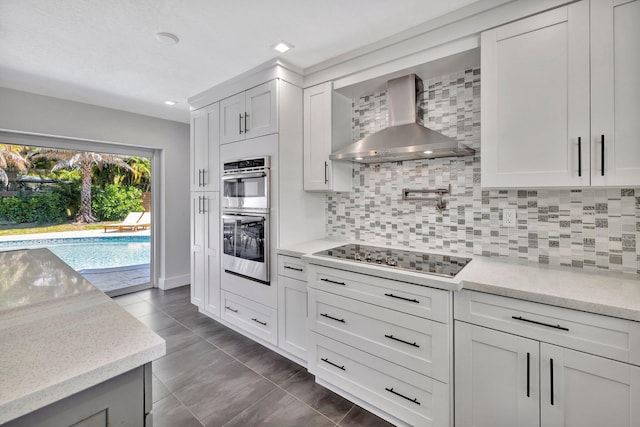 kitchen with black electric stovetop, wall chimney range hood, decorative backsplash, stainless steel double oven, and white cabinets