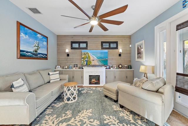 living room featuring wooden walls, visible vents, an accent wall, a premium fireplace, and a ceiling fan